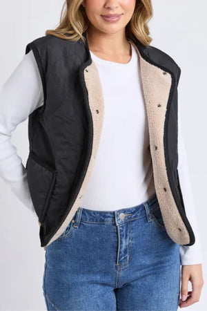 A woman wears the Foxwood Alberta Vest, a black quilted vest with beige sherpa lining, layered over a white long-sleeve shirt and blue jeans. She stands with one arm bent at her side.
