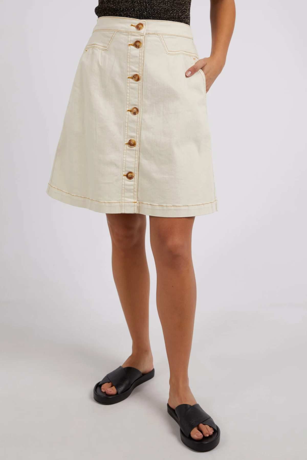 Wearing the Foxwood Amanda Skirt in cream with button-down front and pockets, paired with a dark top and black slide sandals, this look exudes a subtle retro vibe against a plain light background.