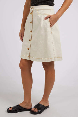 Someone stands against a light background in the cream Foxwood Amanda Skirt, featuring front buttons and side pockets. Black slide sandals complete the subtle retro look, with one hand casually in their pocket.