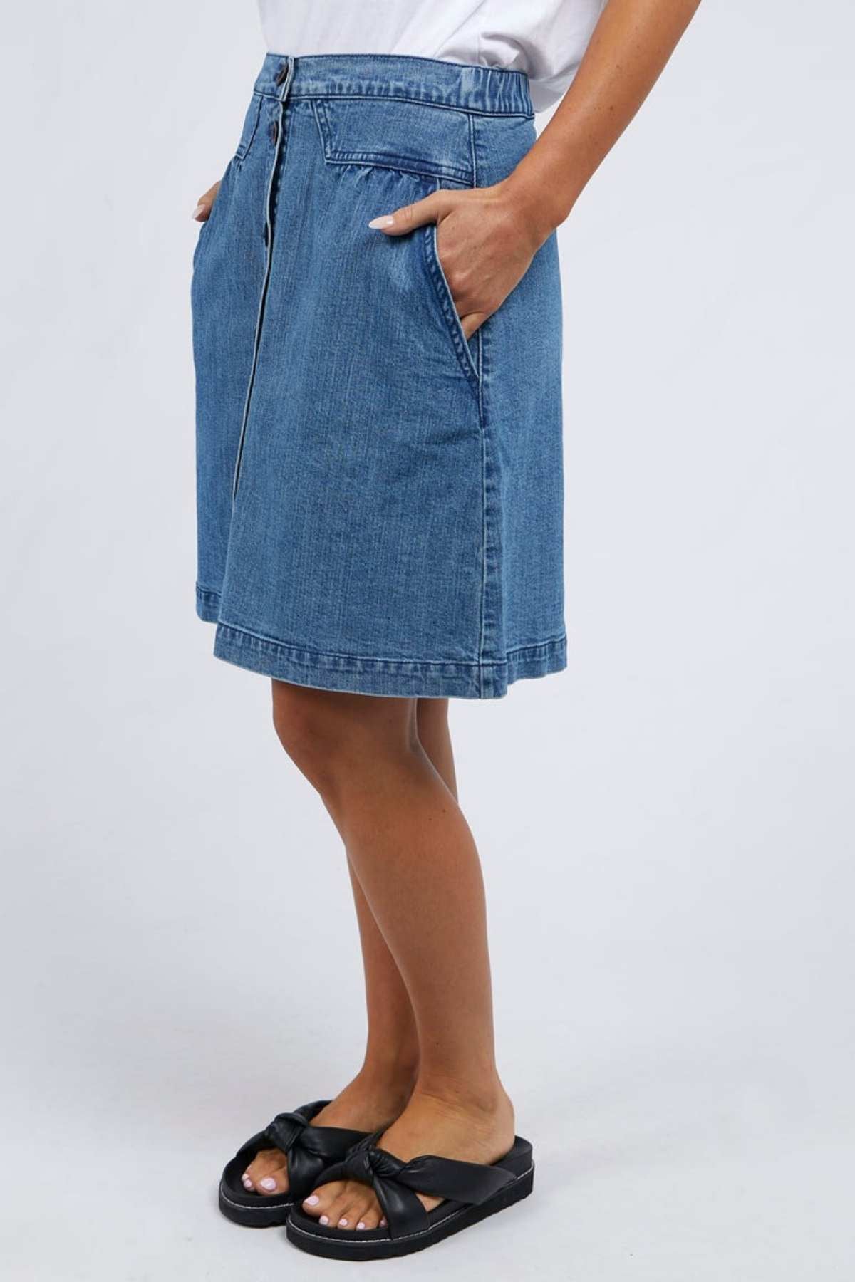 The individual is wearing the Foxwood Amanda Skirt, which falls to knee-length in a stylish blue denim. It's paired with a crisp white top and chic black sandals. With their hands cozily tucked into the skirt's pockets, they exude a retro vibe against a neutral backdrop.