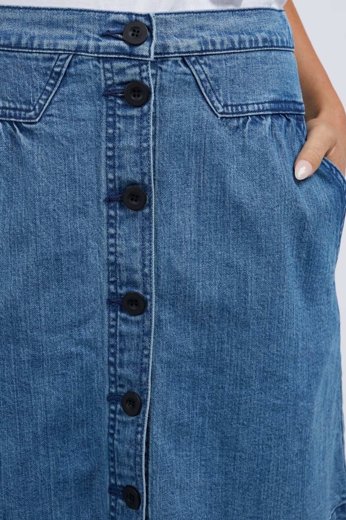 Close-up of a person wearing the Foxwood Amanda Skirt, which features an A-Line cut with front buttons. The skirt exudes a retro vibe, complete with two pockets—one partially visible as a hand is tucked inside—and its classic blue denim texture adds to its timeless appeal.