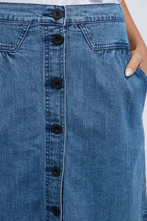 Close-up of a person wearing the Foxwood Amanda Skirt, which features an A-Line cut with front buttons. The skirt exudes a retro vibe, complete with two pockets—one partially visible as a hand is tucked inside—and its classic blue denim texture adds to its timeless appeal.