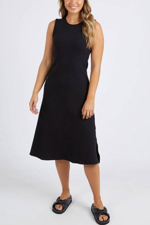 A woman smiles against a plain white background, wearing the Foxwood Amelie Rib Dress and black slide sandals, with one arm relaxed by her side.