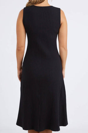 A woman models the Foxwood Amelie Rib Dress—a sleeveless, knee-length black dress made from 100% cotton rib—facing away to show its back against a plain light background.