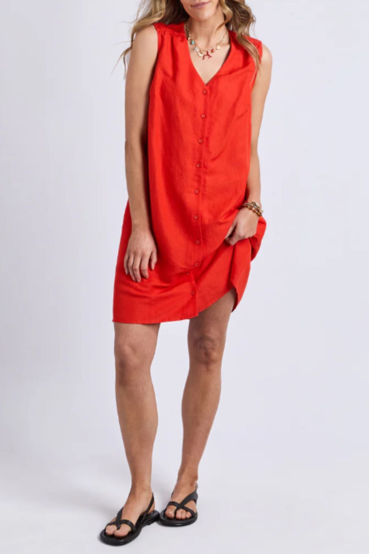 A woman in a sleeveless, button-down Foxwood Anara Dress with a V-neckline stands against a light background, paired with black sandals and a beaded necklace.
