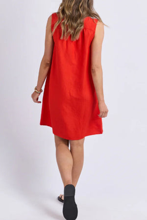A woman with long wavy hair is shown from behind wearing the sleeveless, loose-fitting Foxwood Anara Dress with a V-neckline and black sandals, walking away against a plain light background.