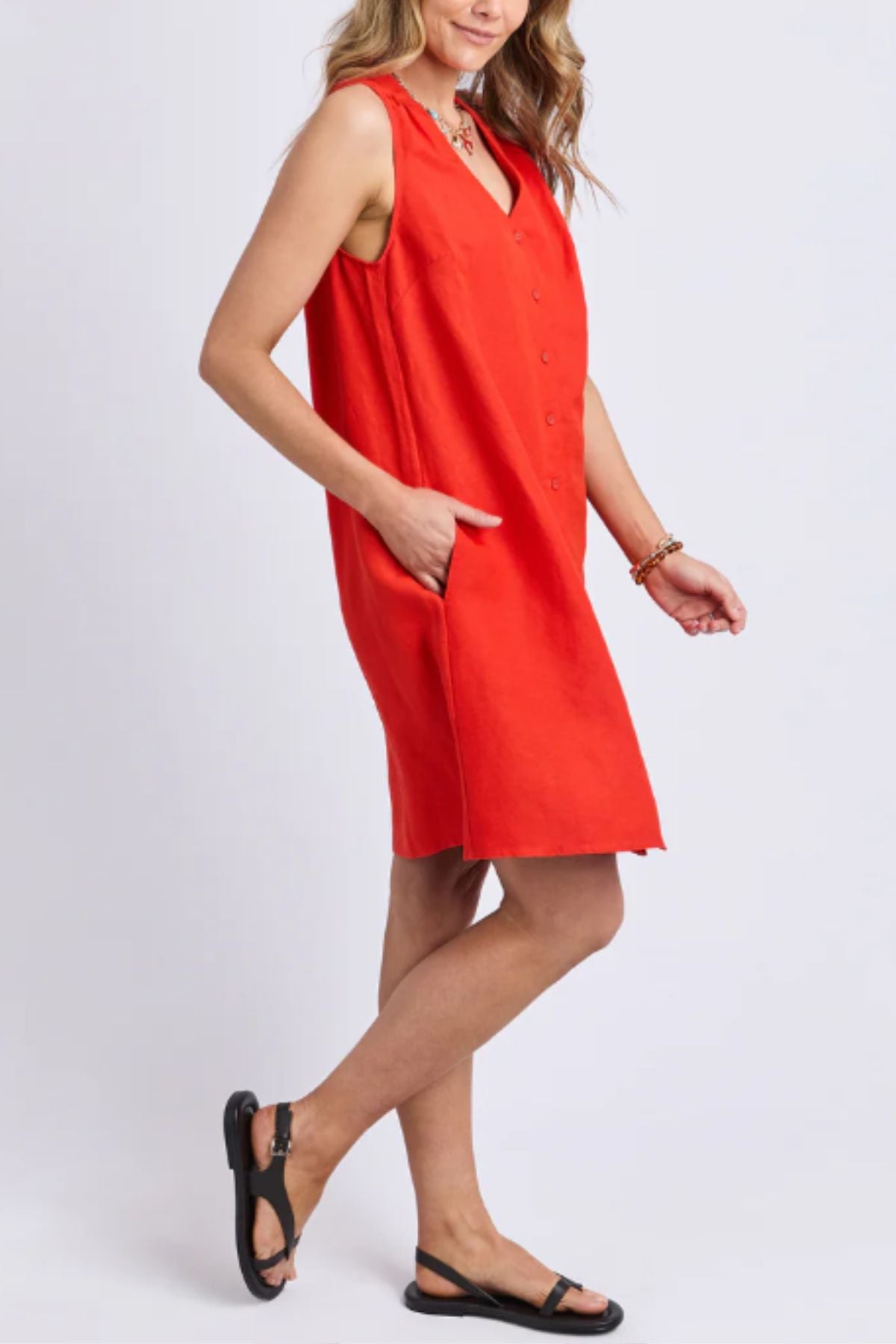 A person stands against a plain light background wearing the Foxwood Anara Dress, a sleeveless, knee-length bright red linen dress with a V-neckline, button details, side pockets, and black sandals.