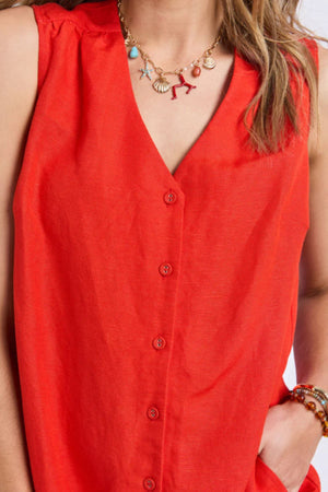 A person wearing the sleeveless, bright red Foxwood Anara Dress with a V-neckline, paired with a beaded bracelet and a gold chain necklace adorned with colorful charms like shells and a starfish.