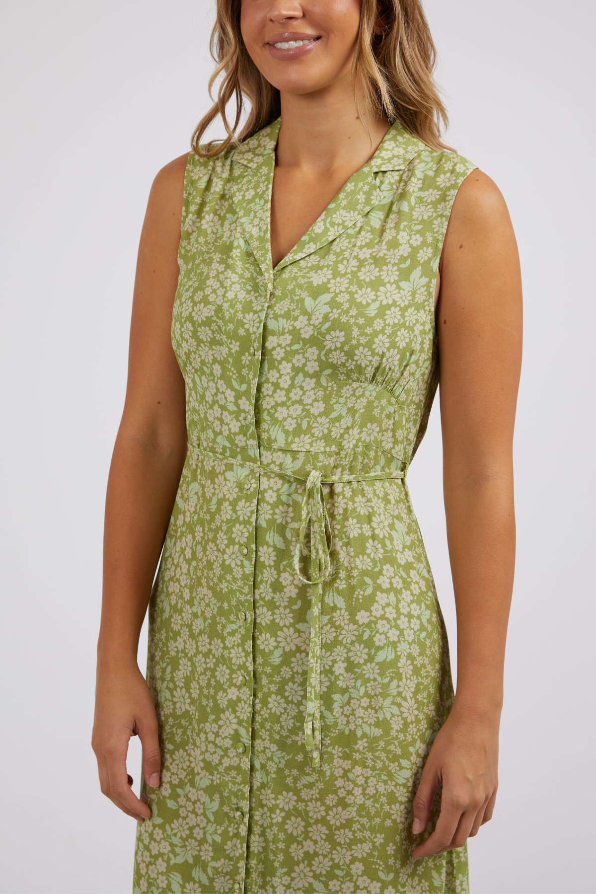 Dressed in the Foxwood Anatayla Floral Dress, a woman exudes elegance with this sleeveless green outfit adorned with florals, ideal for warm weather gatherings. Made from lightweight fabric, it showcases a chic collar and waist tie against a plain white backdrop.