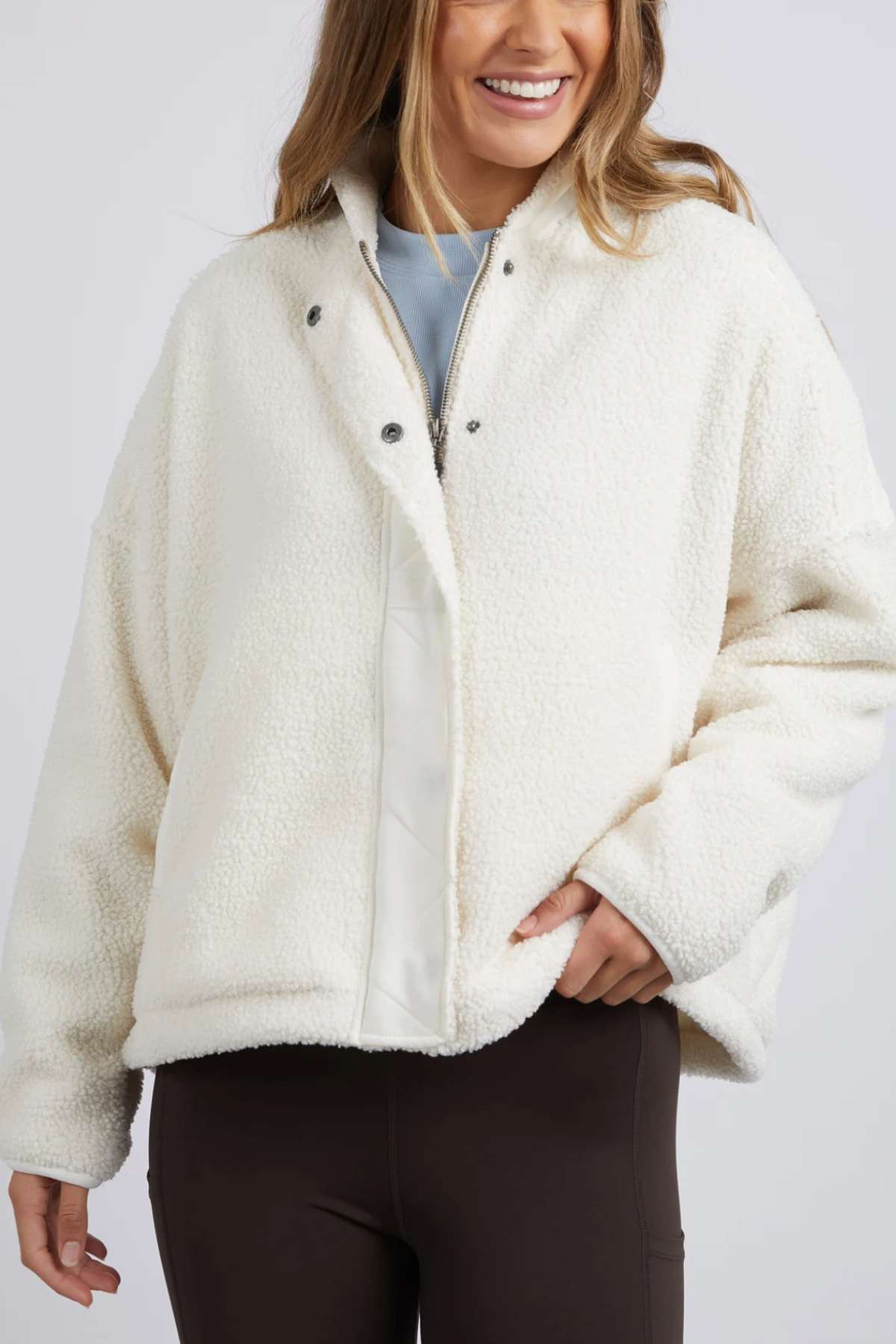A person with long hair, smiling, is wearing a white Foxwood Arizona Fleece Zip Thru jacket with a snap button front and their right hand in one of the jacket's pockets. Theyβre also dressed in brown leggings. The background is plain and light-colored.