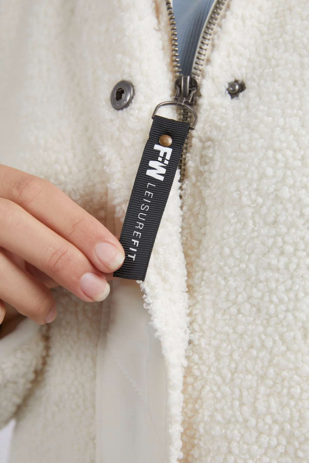 A close-up shot of a person holding a zipper pull tab on a Foxwood Arizona Fleece Zip Thru jacket. The pull tab is black with white text that reads "FW LEISURERIFT." The person is using their left hand, and the zipper is partially open, revealing a light-colored inner liner made of soft teddy fleece.