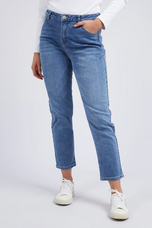 A person in Foxwood Barkly Straight Leg Jeans, a white long-sleeve top, and white sneakers stands with one hand in their pocket against a plain light background.