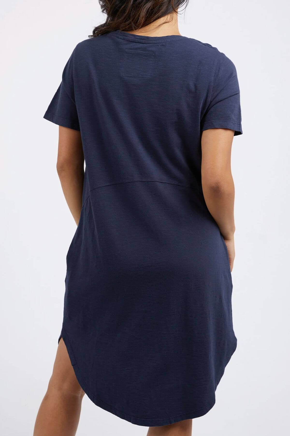 The rear view showcases the elegant fall of the Foxwood Bay Dress, a navy blue short-sleeved jersey dress. Its relaxed fit and cotton comfort make it ideal for any casual wardrobe. The dress falls stylishly above the knee as the person stands against a plain white background.