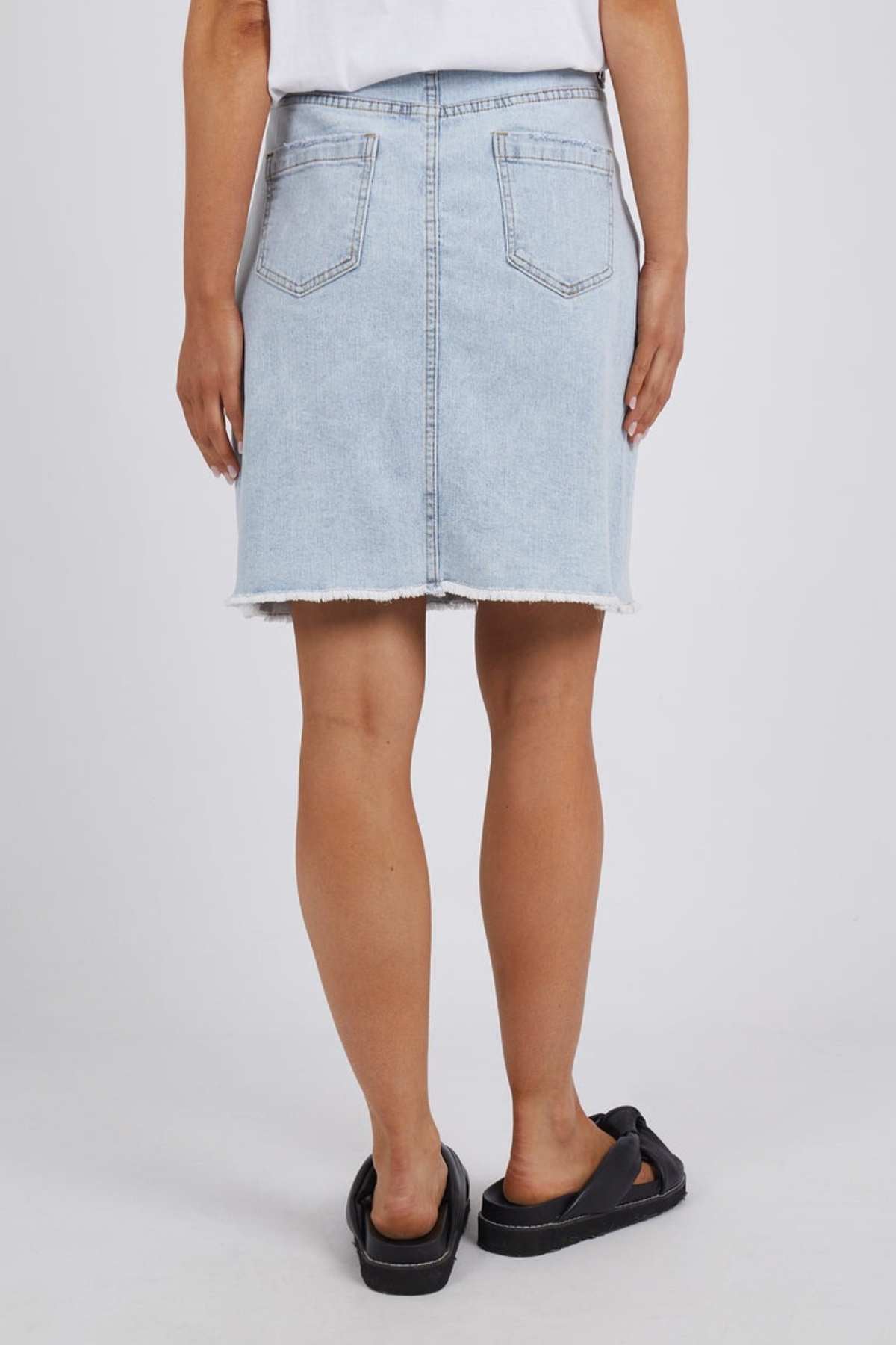 Rear view of a person wearing the light blue Foxwood Belle Skirt, paired with a white top and black sandals. The A-line skirt features a raw hem and back pockets against a neutral gray background.