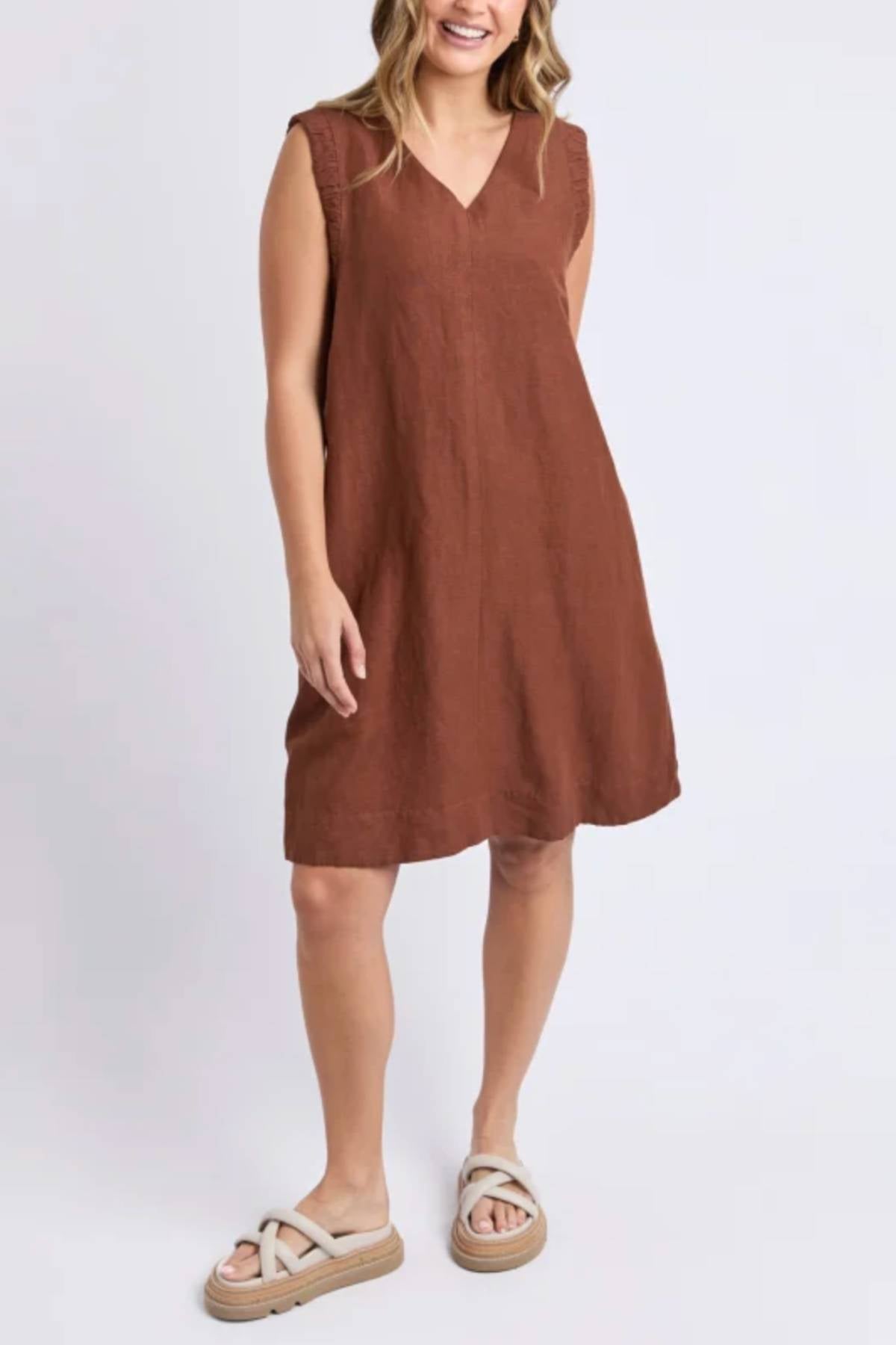 A woman stands against a plain light background, smiling with her hands at her sides, wearing the sleeveless, loose-fitting Foxwood Bermuda Dress in brown and light slip-on sandals.