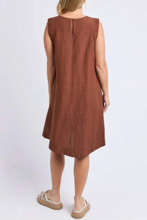 A woman with light skin wears the Foxwood Bermuda Dress—a sleeveless, knee-length brown linen blend dress with a loose fit and ruffled shoulder details. She faces away and pairs it with beige platform sandals.
