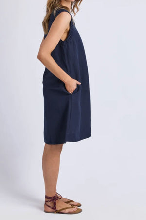 A woman stands in profile against a plain light background, hands in her pockets, wearing the Foxwood Bermuda Dress—a sleeveless, loose-fitting navy linen-blend design—paired with brown flat ankle-strap sandals.