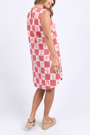 A woman smiles sideways in the Foxwood Biarittz Dress—a sleeveless, knee-length cotton shift in a red and white checkerboard pattern—paired with beige wedge sandals against a plain light background.