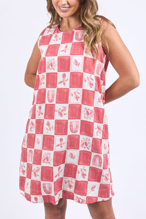 A woman in a sleeveless, knee-length Foxwood Biarittz Dress with red and white checks, floral and fruit patterns, stands against a plain light background and smiles.