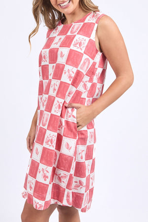 A woman smiles with her hand in the pocket of a sleeveless Foxwood Biarittz Dress, featuring a red and white checkered pattern with floral and fruit designs, against a plain light background.