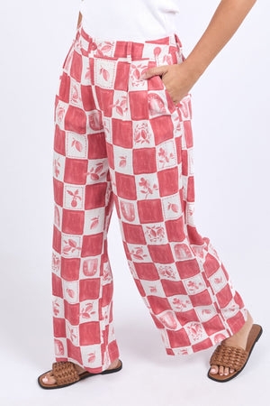 Wearing loose-fitting Foxwood Biarittz Pant with a red and white checkered floral pattern, one hand in the pocket, paired with a white top and brown woven sandals. Made from a soft linen-viscose blend for comfort all day.
