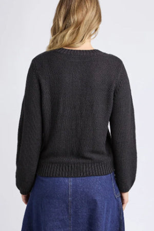 A woman with wavy blonde hair is seen from behind, wearing the Foxwood Blair Long Sleeve Knit in black and a blue denim skirt against a plain light gray background.