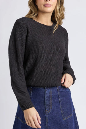 A woman wears the Foxwood Blair Long Sleeve Knit in black with a high-waisted blue denim skirt, standing against a light background, left arm bent and right hand at her side.