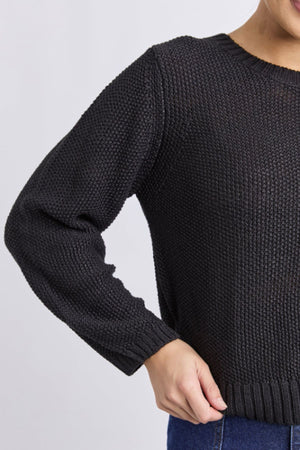 A person wears the Foxwood Blair Long Sleeve Knit, a relaxed-fit black cotton sweater, paired with blue jeans. They stand with one hand on their hip against a plain light background, showing only the upper body and part of the face.
