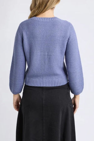 A person with light brown hair is shown from the back, wearing a Foxwood Blair Long Sleeve Knit in lavender and a black skirt. The knit has a relaxed fit and long sleeves, set against a plain, light-colored background.