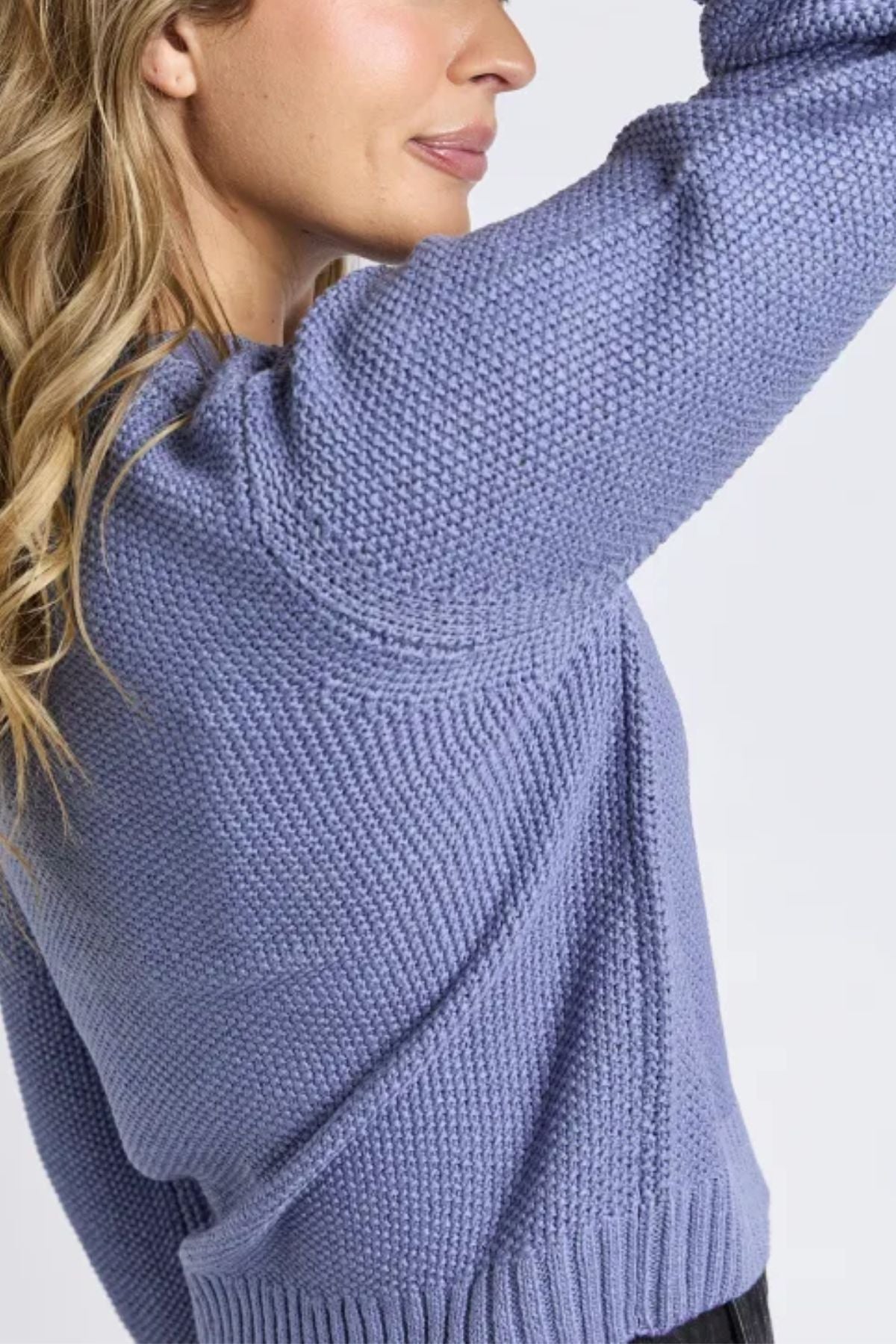 A woman with long, wavy blonde hair wears the Foxwood Blair Long Sleeve Knit in light purple. Only her lower face and torso are shown as she lifts one arm, partially covering her face.