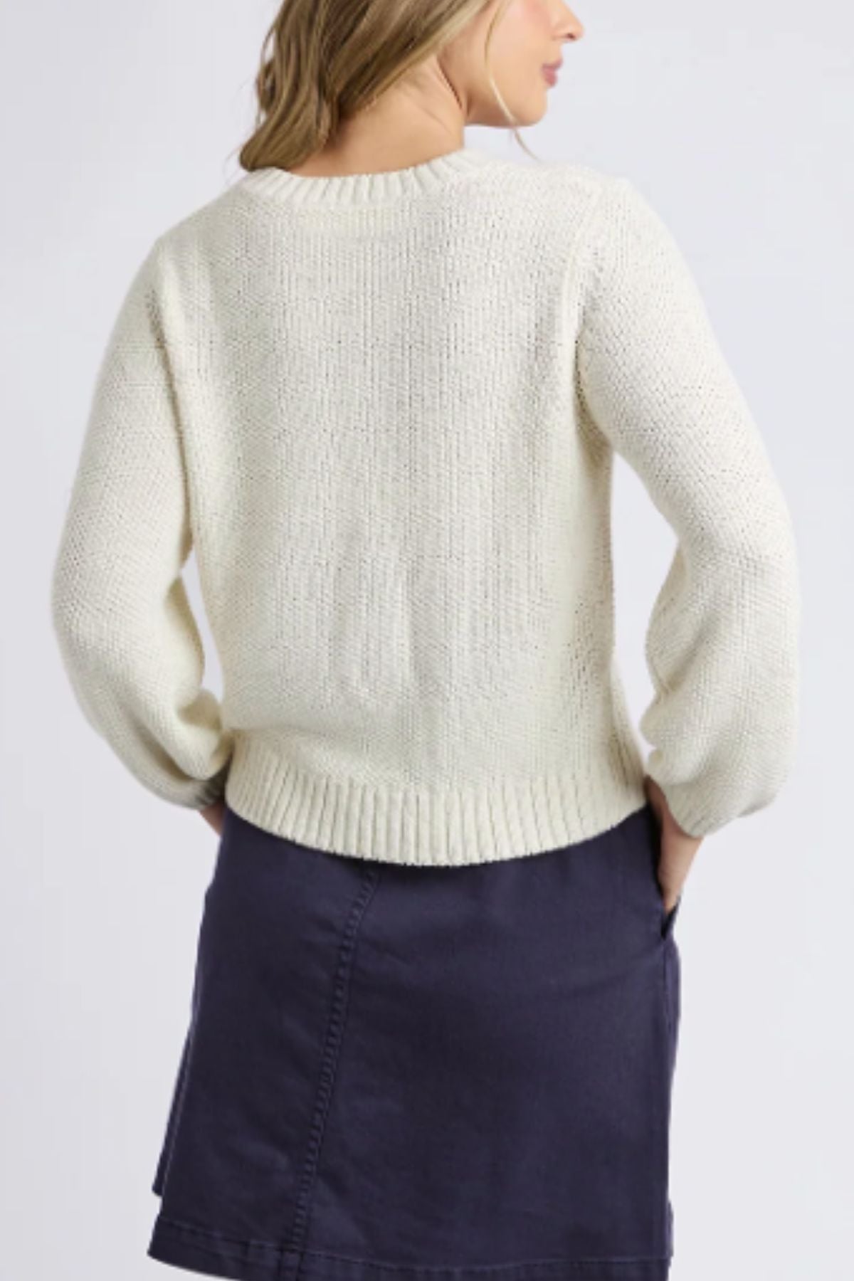 A woman faces away from the camera, wearing the Foxwood Blair Long Sleeve Knit in cream and a navy blue skirt. Her hands rest in her skirt pockets, and her light brown hair cascades over her shoulders.