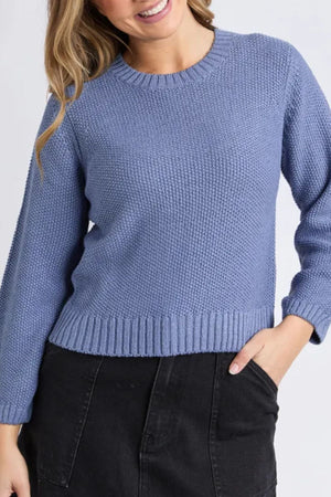 A woman wears the Foxwood Blair Long Sleeve Knit, a light blue relaxed-fit sweater made from breathable fabric, paired with a black skirt. She smiles with one hand in her skirt pocket against a plain, light background.