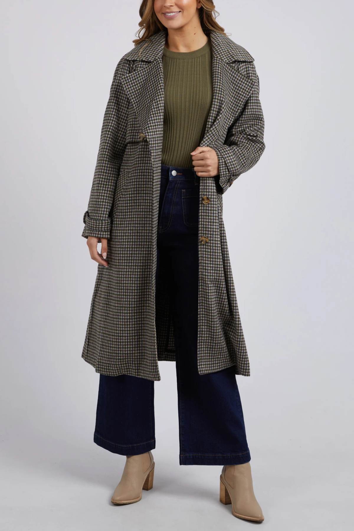 A person is wearing a green ribbed sweater, dark blue wide-leg jeans, and the Foxwood Brodie Trench coat—a long plaid piece made from a wool polyester blend with front welt pockets. They are also wearing beige ankle boots. The person is standing against a plain, light gray background.