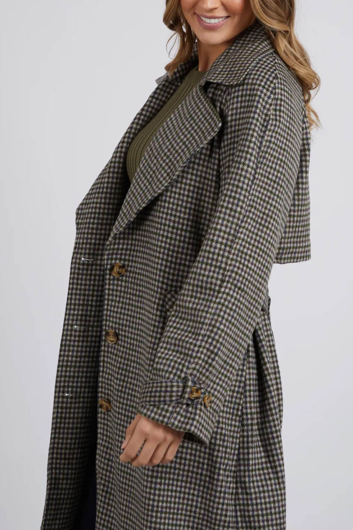 A woman is wearing a Foxwood Brodie Trench, a long, checkered coat with large buttons and front welt pockets. The wool polyester blend coat features a broad collar and extended cuffs. She has wavy, shoulder-length hair and is smiling against the plain white background.