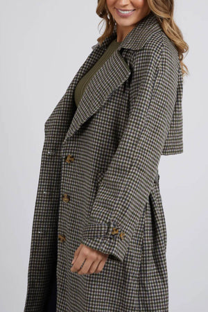 A woman is wearing a Foxwood Brodie Trench, a long, checkered coat with large buttons and front welt pockets. The wool polyester blend coat features a broad collar and extended cuffs. She has wavy, shoulder-length hair and is smiling against the plain white background.