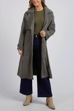 A person is wearing a green ribbed sweater, dark blue wide-leg jeans, and the Foxwood Brodie Trench coat—a long plaid piece made from a wool polyester blend with front welt pockets. They are also wearing beige ankle boots. The person is standing against a plain, light gray background.
