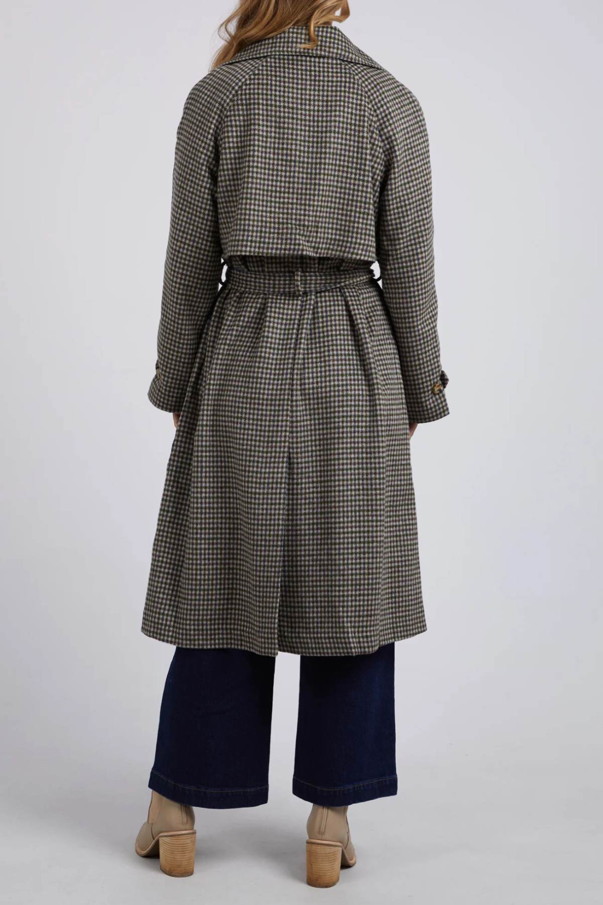 A person with long hair is seen from behind, wearing the Foxwood Brodie Trench crafted from a chic wool polyester blend, featuring front welt pockets. They are also dressed in wide-leg dark jeans and beige heeled shoes, set against a plain light-colored background.
