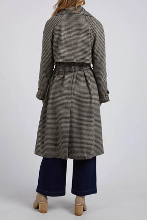 A person with long hair is seen from behind, wearing the Foxwood Brodie Trench crafted from a chic wool polyester blend, featuring front welt pockets. They are also dressed in wide-leg dark jeans and beige heeled shoes, set against a plain light-colored background.