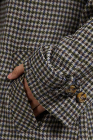 A detailed view showcases an individual wearing the Foxwood Brodie Trench with a hand tucked into the side pocket. The trench boasts a striking pattern of green, blue, and beige checks, complemented by brown buttons on the cuff. Made from a blend of wool and polyester, it promises both comfort and durability.