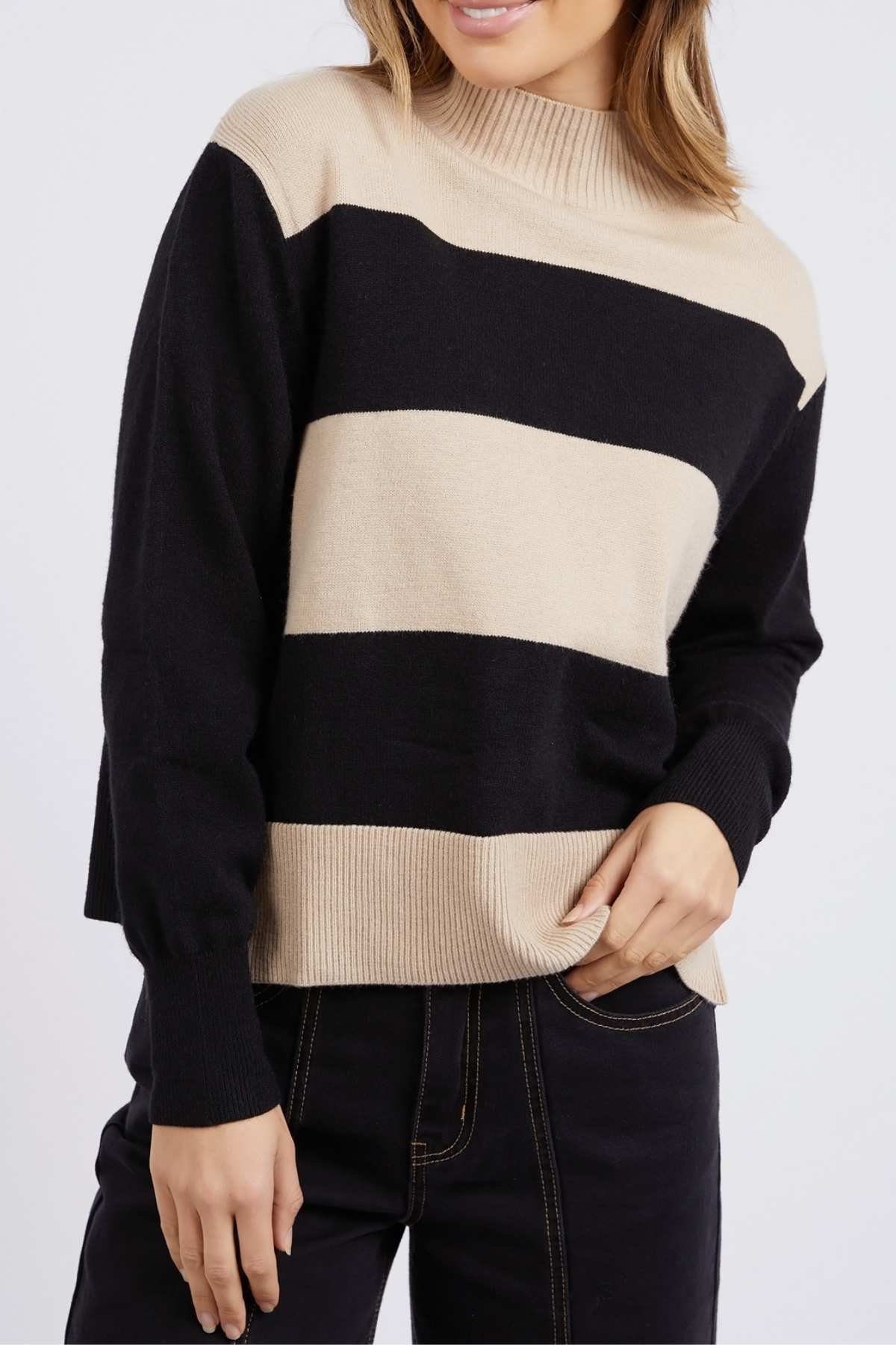The individual sports the Foxwood Canterbury Knit, a relaxed fit sweater with beige and black wide horizontal stripes, paired with dark pants. They're casually posed, one hand tucked into their pocket.