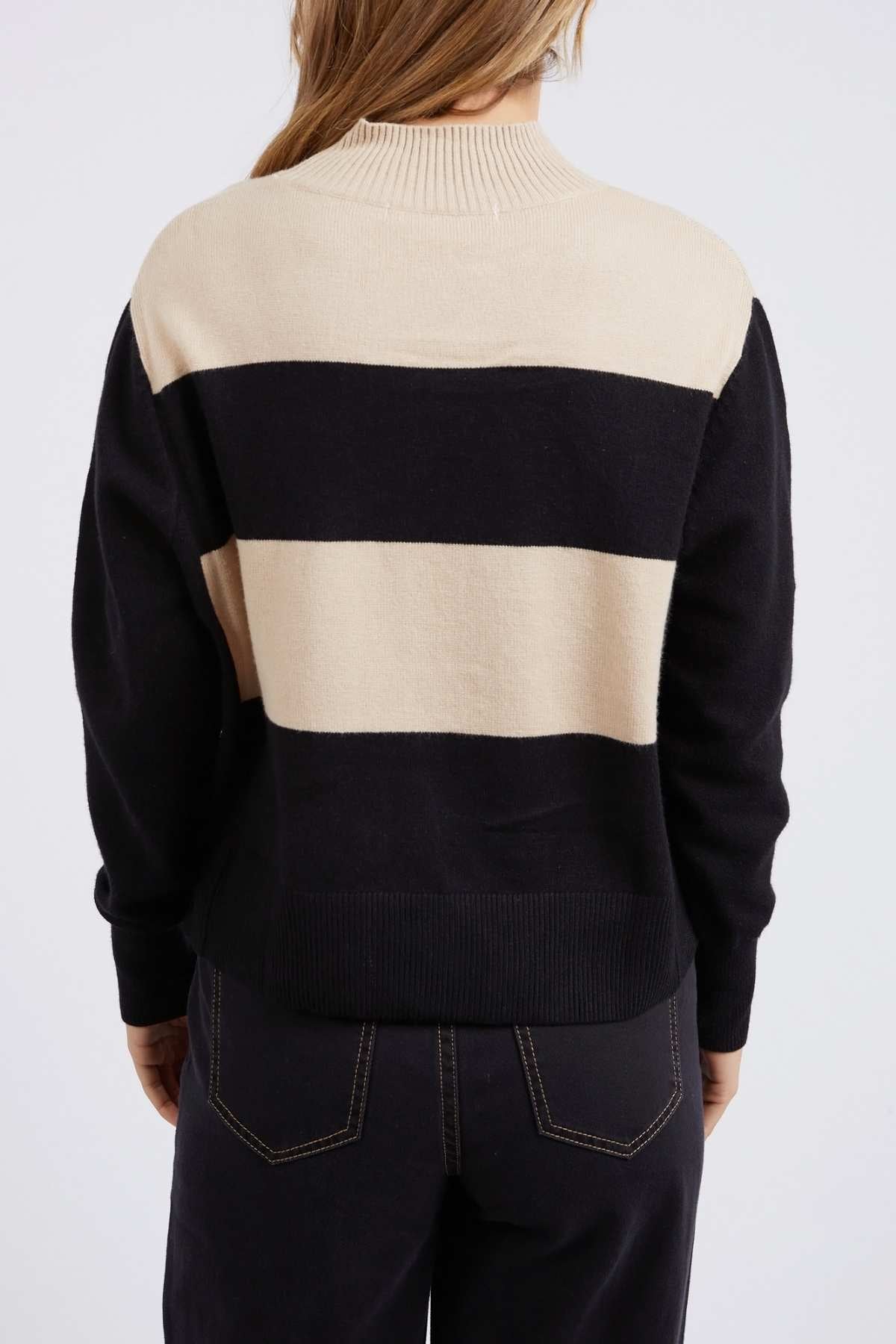 The person with long hair is shown from the back, wearing a Foxwood Canterbury Knit, a relaxed fit sweater with black and beige stripes, paired with dark pants. This soft wool/cashmere blend has ribbed collar and cuffs over a plain white backdrop.