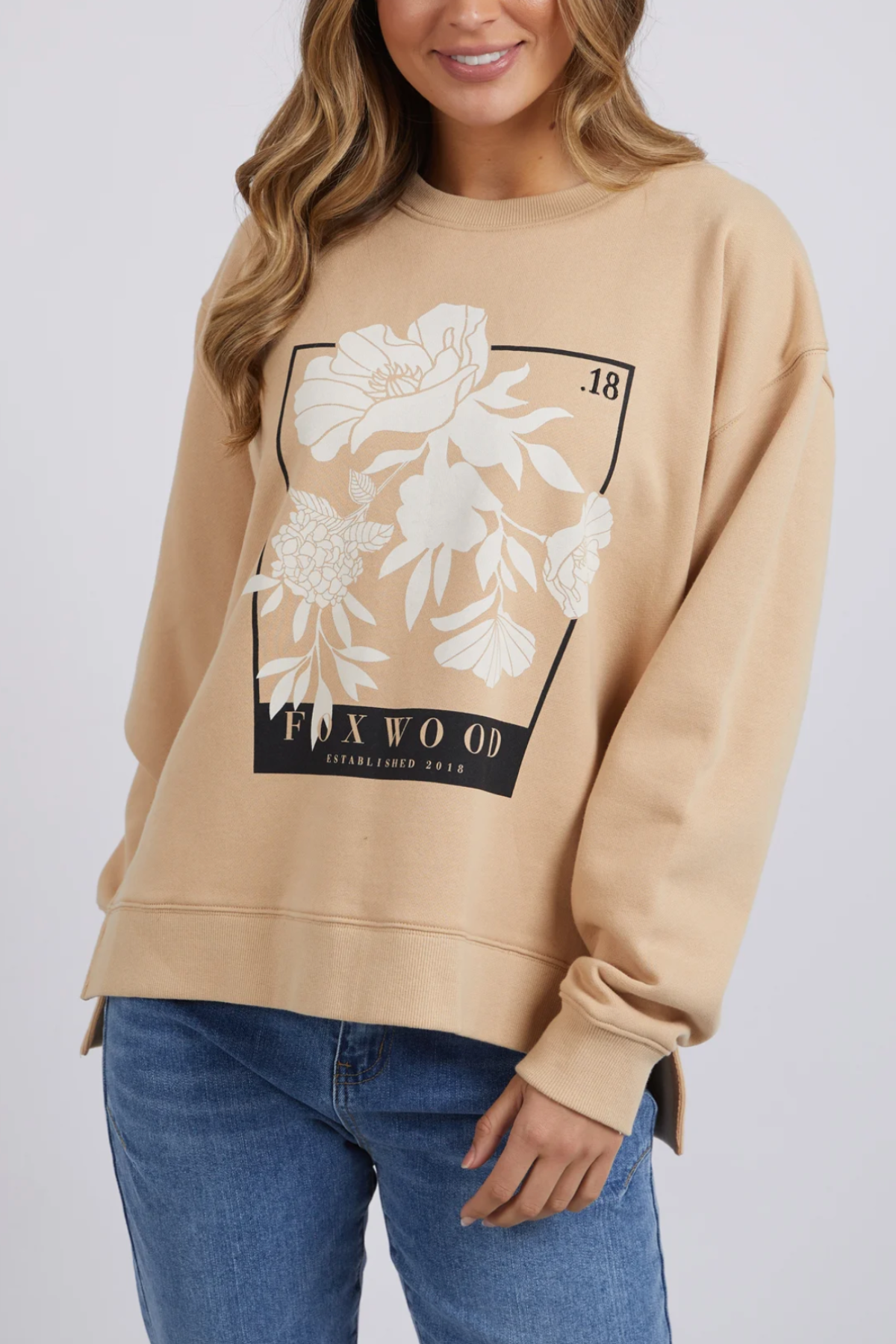 A person with long hair is wearing the Foxwood Carnation Crew, a relaxed-fit jumper crafted from 100% cotton. The beige sweater showcases a floral design along with the words "FOXWOOD" and "Established 2014" on the front. They are also wearing blue jeans and standing against a plain background.