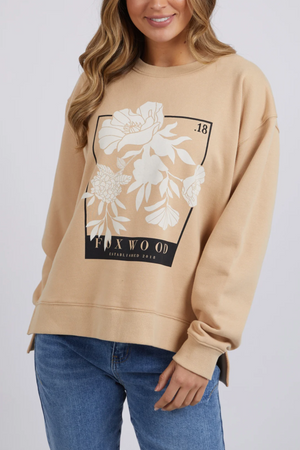 A person with long hair is wearing the Foxwood Carnation Crew, a relaxed-fit jumper crafted from 100% cotton. The beige sweater showcases a floral design along with the words "FOXWOOD" and "Established 2014" on the front. They are also wearing blue jeans and standing against a plain background.