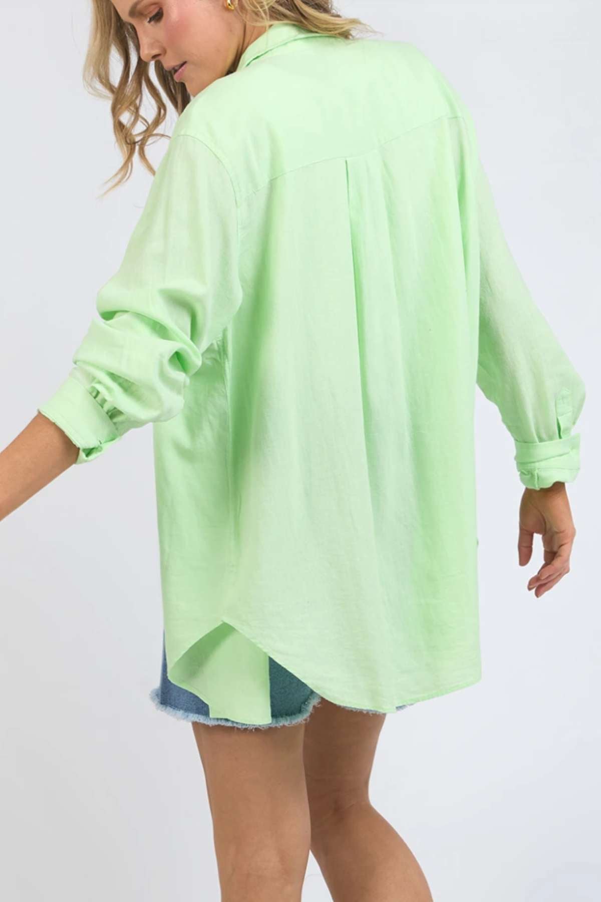 A person wearing a light green Foxwood Carrie Shirt with rolled-up sleeves and light blue denim shorts stands facing slightly away from the camera, showcasing its back. The lightweight fabric and relaxed fit of the shirt are evident against the plain, light-colored background.