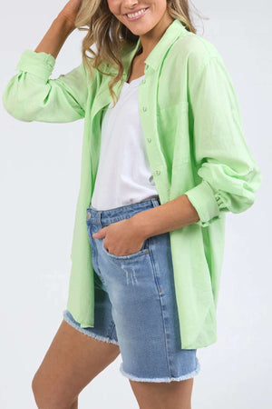 A person dressed in a light green relaxed fit Foxwood Carrie Shirt over a white top, paired with blue denim shorts. The sleeves of the lightweight shirt are rolled up. One hand is bent, touching their head, while the other hand rests in their pocket.