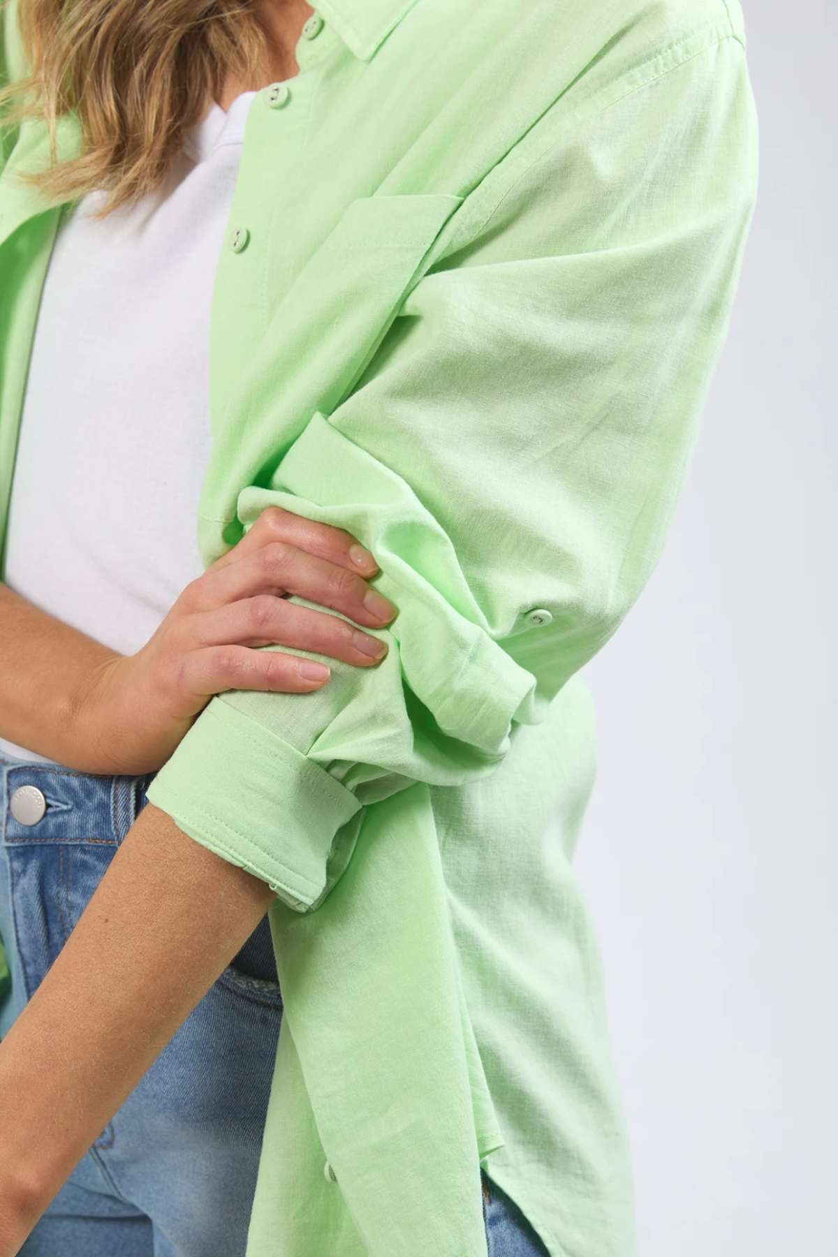 A person with light-colored hair rolls up the sleeves of a light green Foxwood Carrie Shirt, made from lightweight fabric. They are wearing a white t-shirt underneath and blue jeans. The image is a close-up, emphasizing the relaxed fit around the upper body and arms.