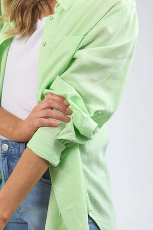 A person with light-colored hair rolls up the sleeves of a light green Foxwood Carrie Shirt, made from lightweight fabric. They are wearing a white t-shirt underneath and blue jeans. The image is a close-up, emphasizing the relaxed fit around the upper body and arms.