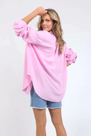 A woman with wavy blonde hair poses in comfort and style, her right hand touching her head. She wears a loose, long-sleeved Foxwood Carrie Shirt in pink paired with relaxed-fit blue denim shorts. The background is plain white.