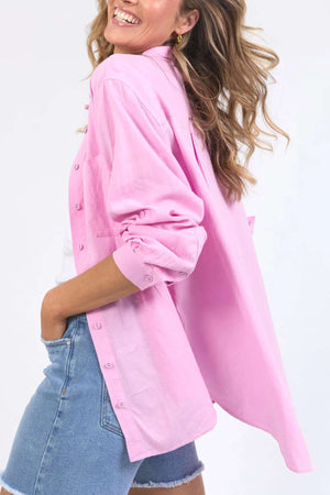 A smiling person with wavy, light brown hair is wearing a Foxwood Carrie Shirt and blue denim shorts. The relaxed-fit, light pink shirt's sleeves are rolled up, billowing as the person strikes a casual and joyful pose with one hand in their pocket, embodying comfort and style.