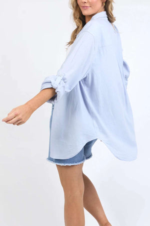 A person with long, wavy hair is seen from the side wearing a light blue Foxwood Carrie Shirt with rolled-up sleeves and denim shorts. The oversized shirt, crafted from breathable fabric, is unbuttoned at the bottom and drapes loosely in its relaxed fit as they extend one arm slightly backward.